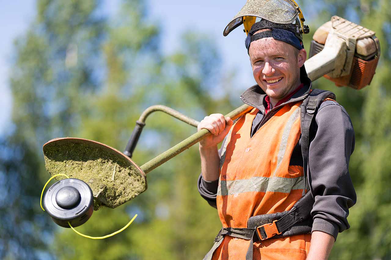 Lächelnder Gärtner in Schutzkleidung hält einen Rasentrimmer, Arbeitsgerät und -umfeld zeigen professionelle Gartenpflege.