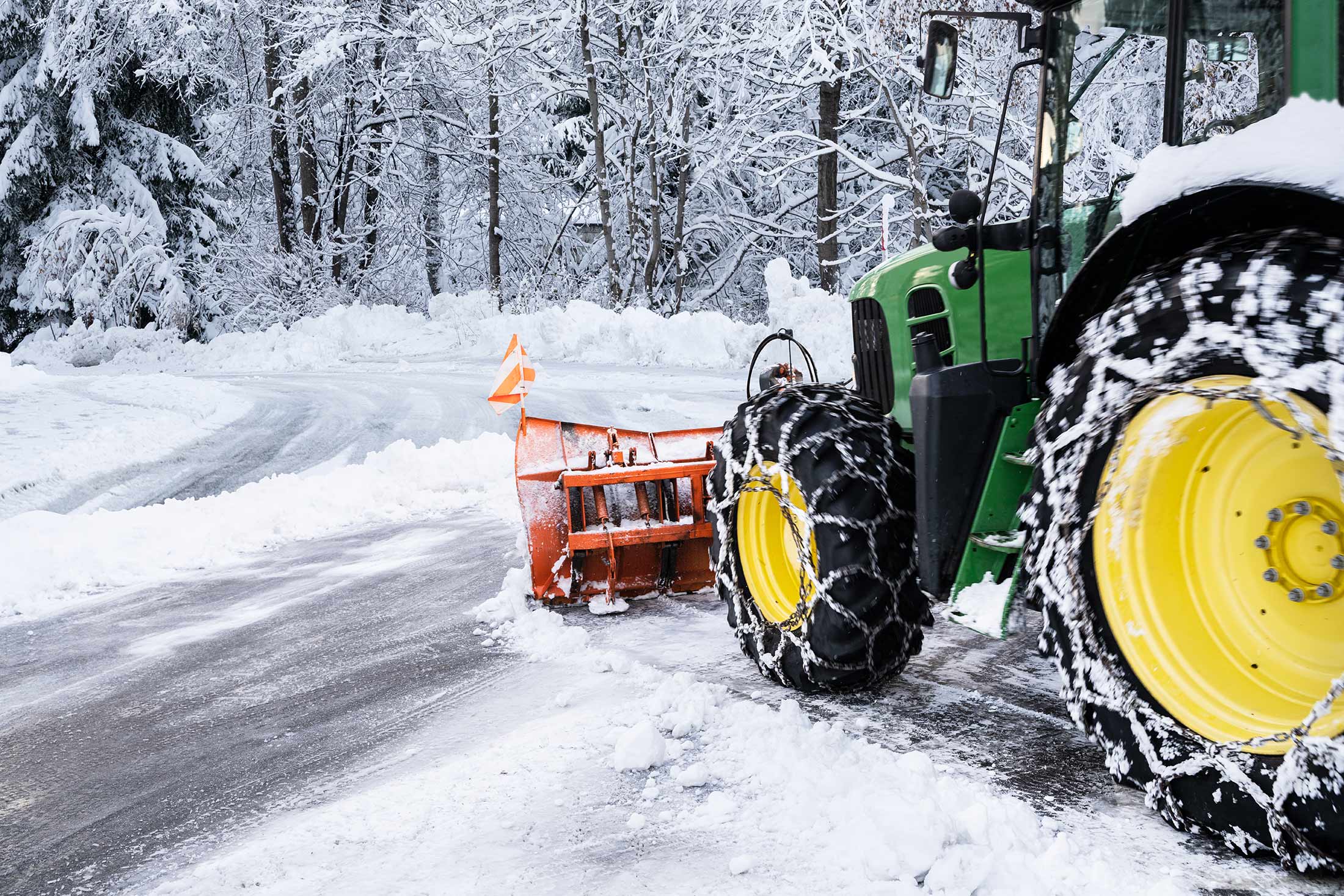 Traktor mit Schneepflug und Ketten räumt Schnee auf einer verschneiten Straße umgeben von winterlichen Bäumen