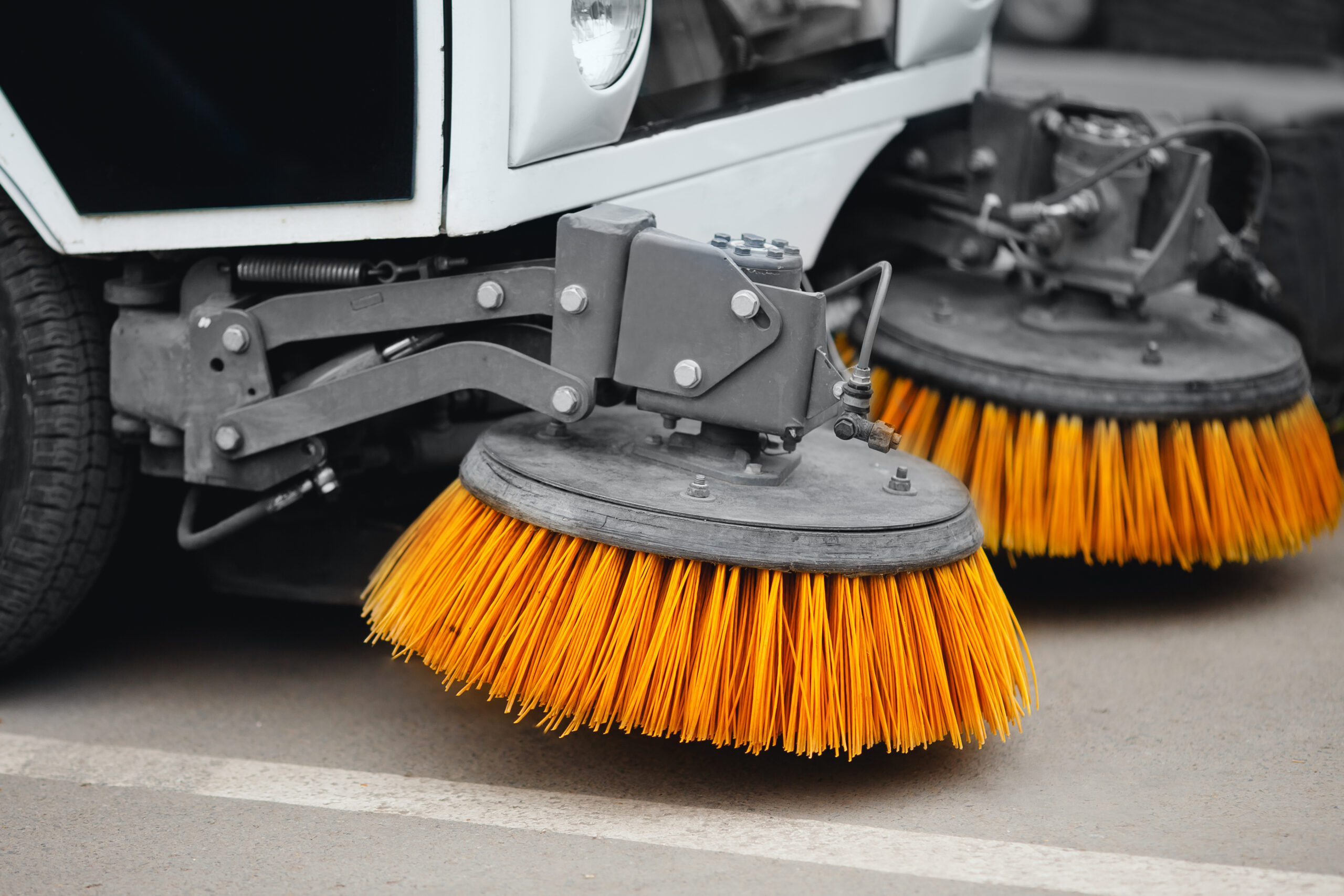 Nahaufnahme einer Kehrmaschine mit rotierenden gelben Bürsten, die eine Asphaltfläche reinigt.